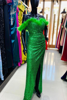 Abito lungo aderente verde in paillettes con scollo a barchetta, piume decorative e spacco laterale, da Moda-Mania.