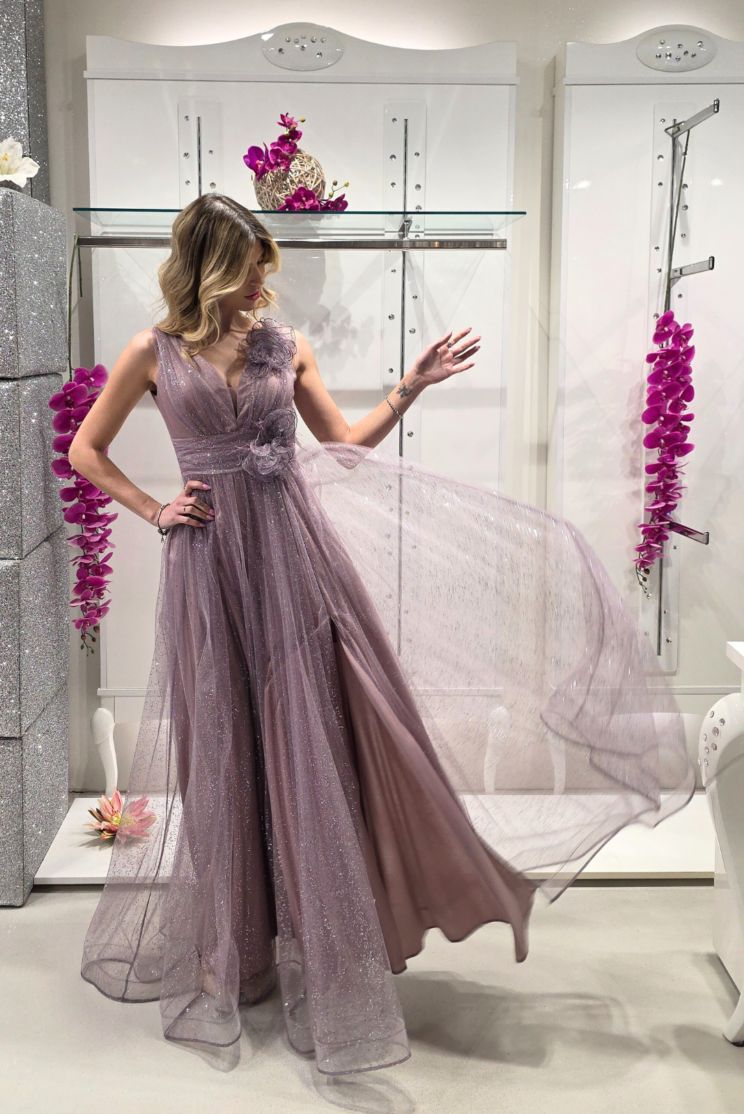Abito lungo svasato lilla in tulle glitterato con spacco e decorazioni floreali, di Moda-Mania, dettaglio del vestito elegante.