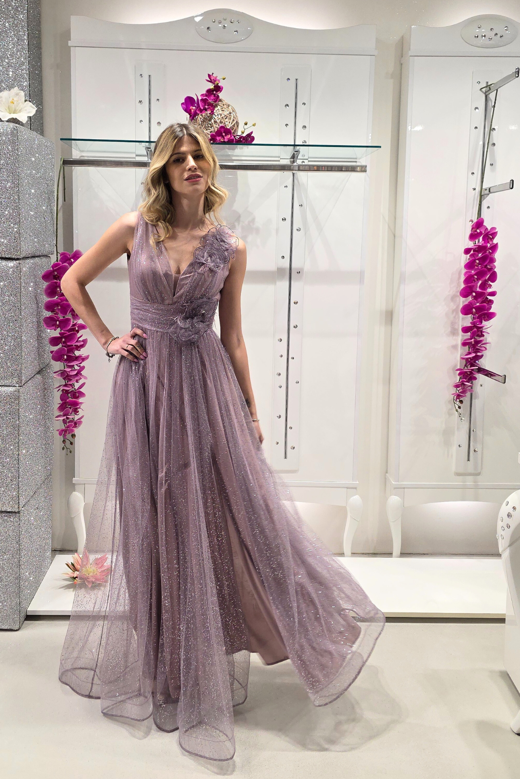 Abito lungo svasato lilla in tulle glitterato con spacco e fiori decorativi, vestito elegante Moda-Mania.