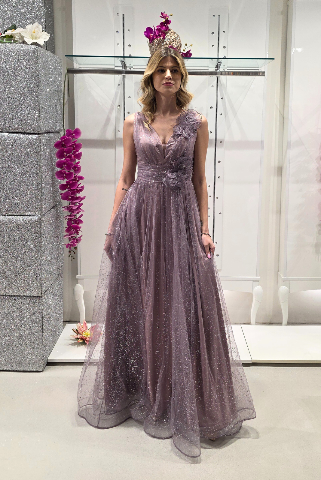 Abito lungo svasato lilla in tulle glitterato con spacco e decorazioni floreali, ideale per occasioni eleganti.