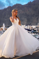 Abito lungo bianco da cerimonia Moda-Mania con corpetto ricamato e gonna ampia in organza su sfondo rosa cipria.
