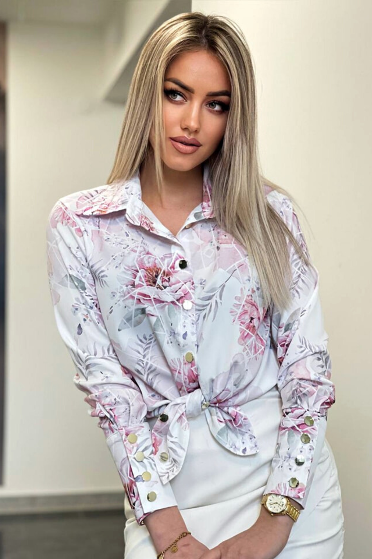 Camicia elegante bianca con fantasia floreale rosa e bottoni dorati, marca Moda-Mania, vista frontale su tessuto leggero.