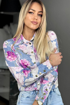 Camicia elegante bianca Moda-Mania con fantasia floreale rosa e bottoni dorati, tessuto leggero e dettagli raffinati.