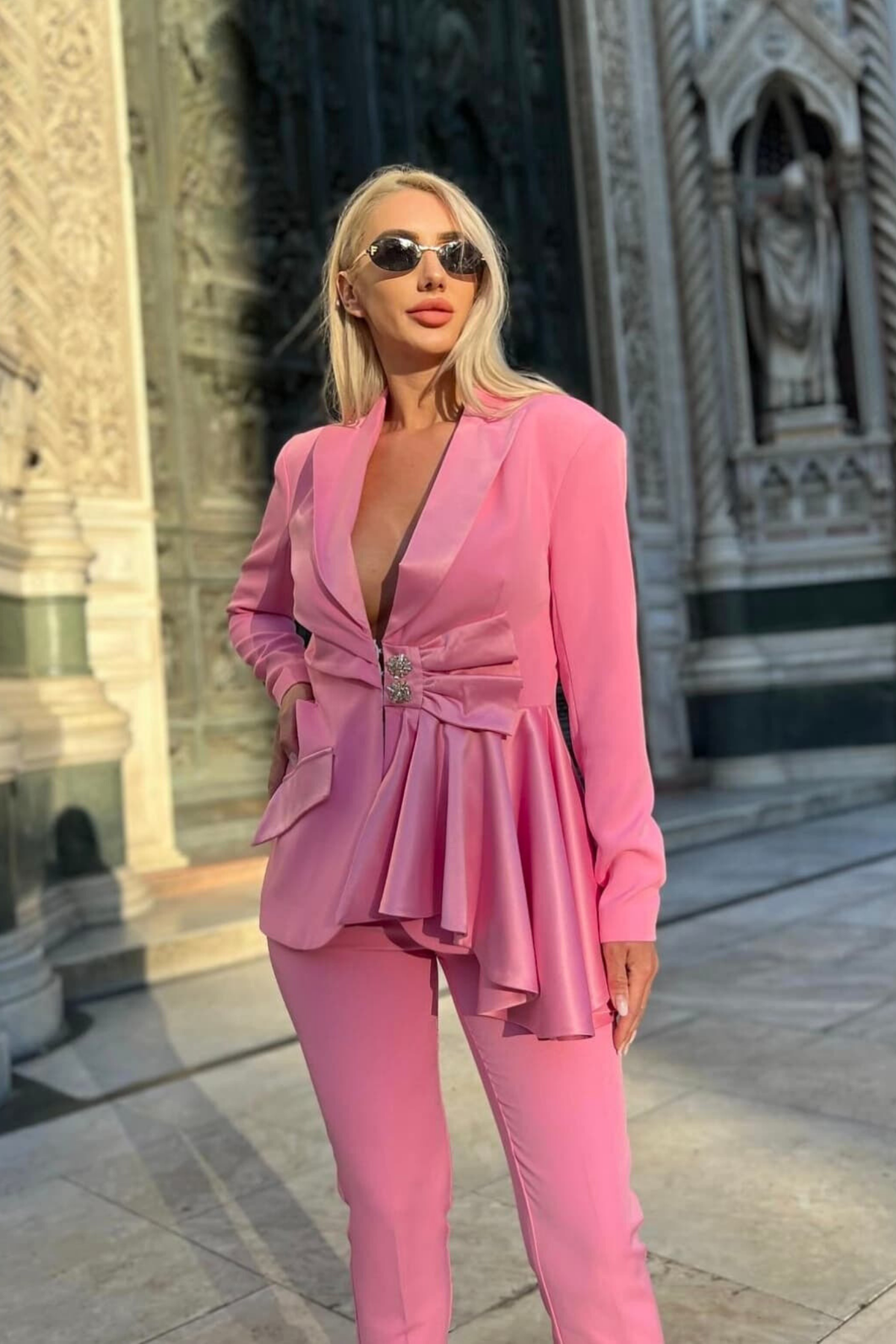 Completo tailleur rosa Bby con giacca e pantaloni, bottoni gioiello, volant, fiocco e cut-out in vita, vestibilità elegante e femminile.