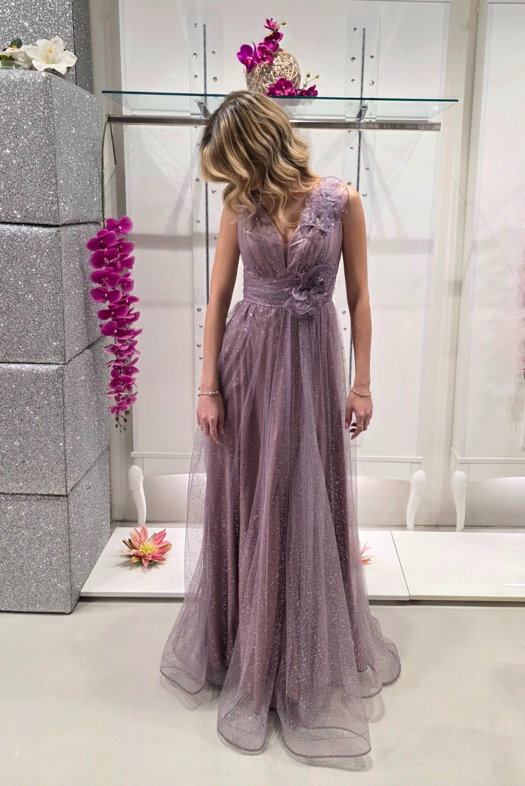 Abito lungo svasato lilla in tulle glitterato con spacco e decorazioni floreali, firmato Moda-Mania, visibile in primo piano.