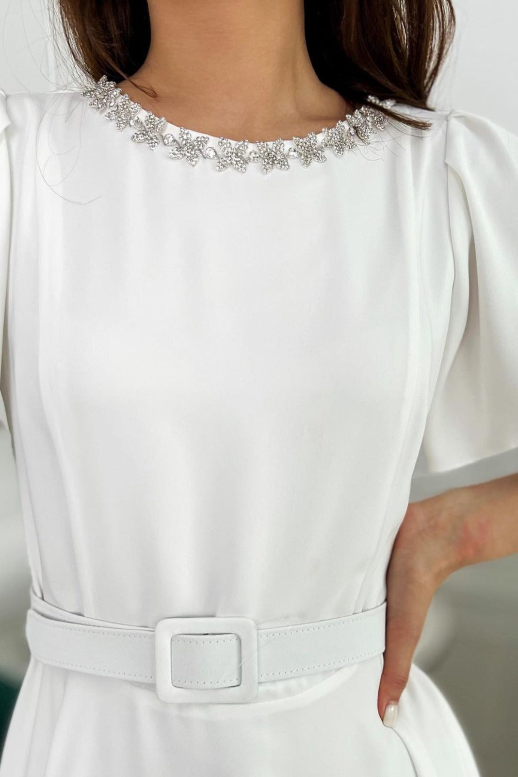 Abito midi svasato bianco Moda-Mania con cintura in vita, tasche laterali e accessorio in strass argento, visto frontalmente.